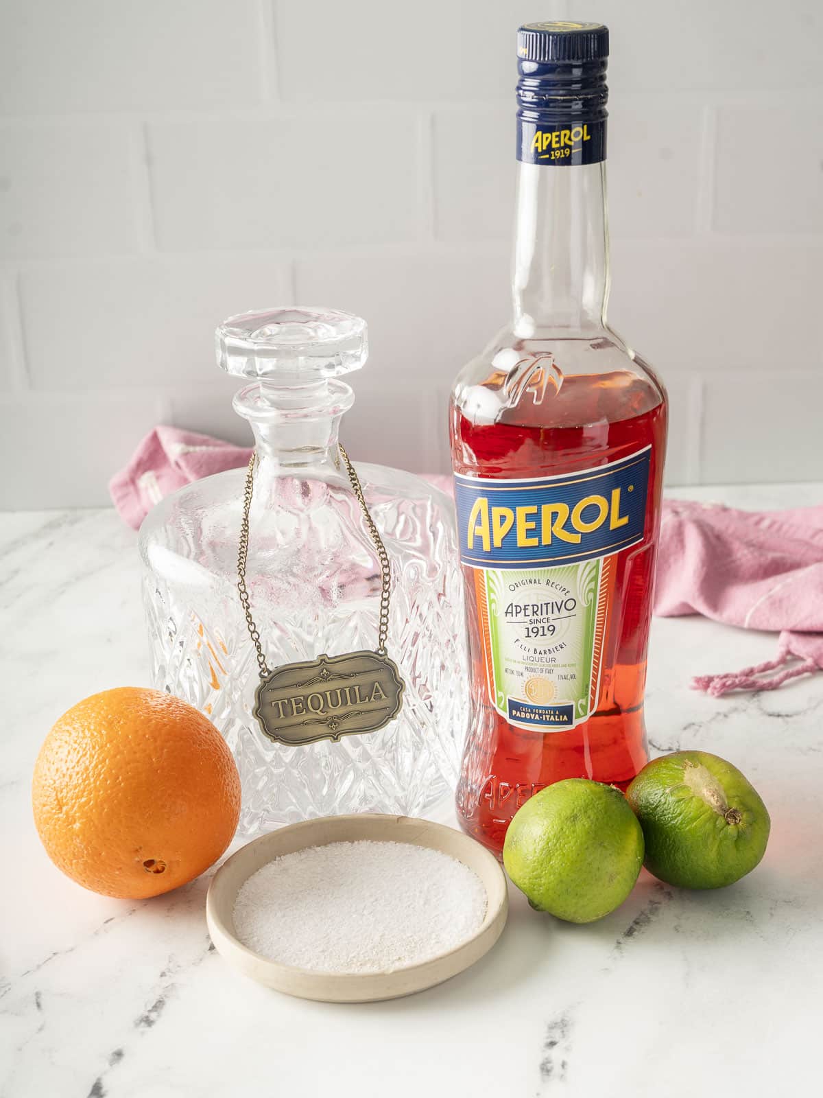 Aperol bottle, tequila decanter, orange, lime, and a bowl of salt rest on a marble surface with a pink cloth, inviting you to craft the perfect Margarita.