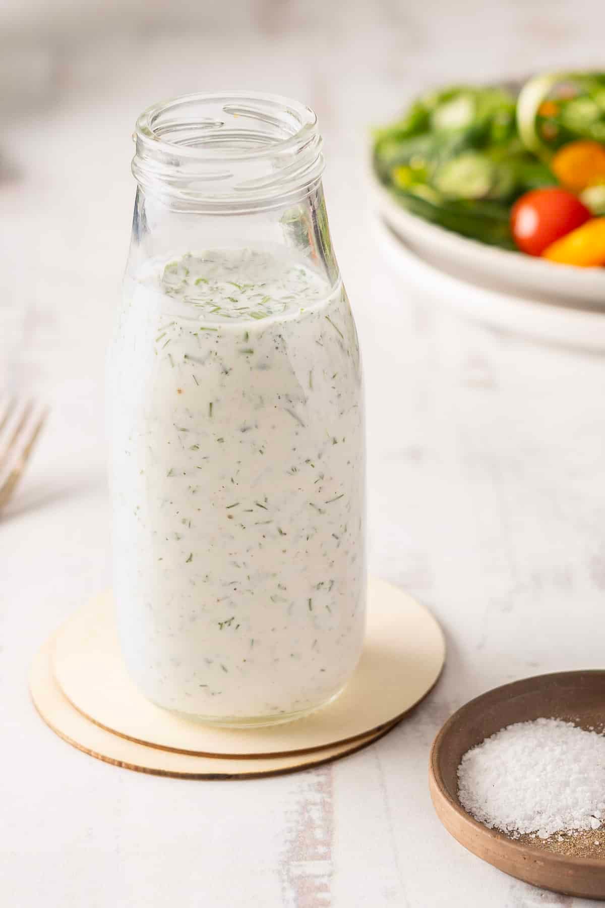 A bottle of creamy buttermilk ranch dressing sits on a coaster, accompanied by a fresh salad and a bowl of salt nearby.