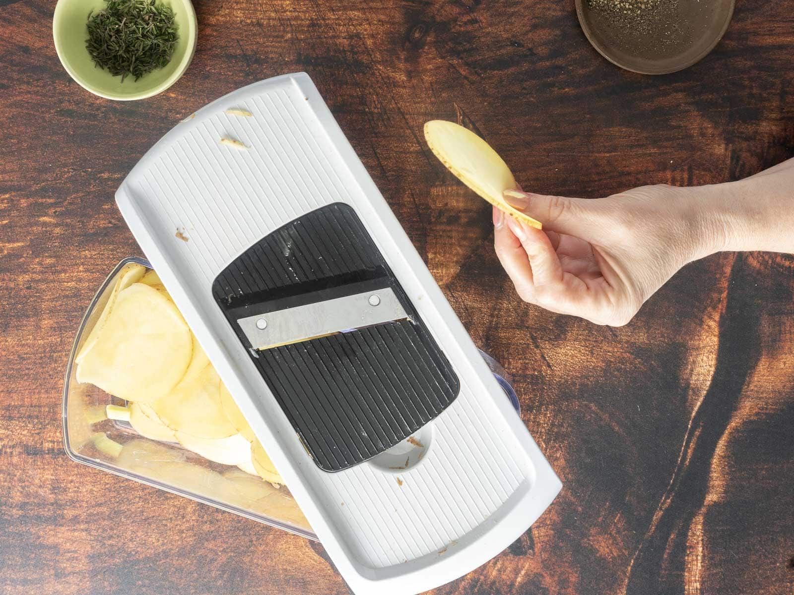 A person holds a potato slice over a mandoline slicer on a wooden surface, preparing for a creamy gratin. 