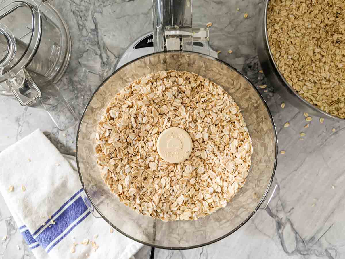 Food processor filled with oats, on a marble countertop beside a cloth towel and a bowl of oats.