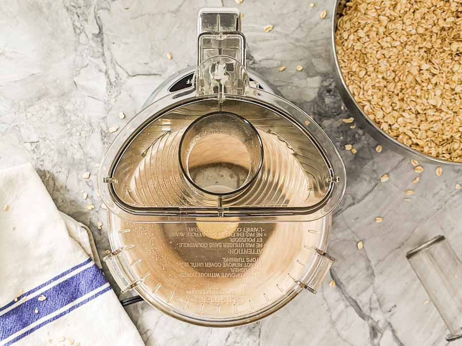 Top view of a food processor bowl next to a bowl of oats on a marble countertop with a kitchen towel.