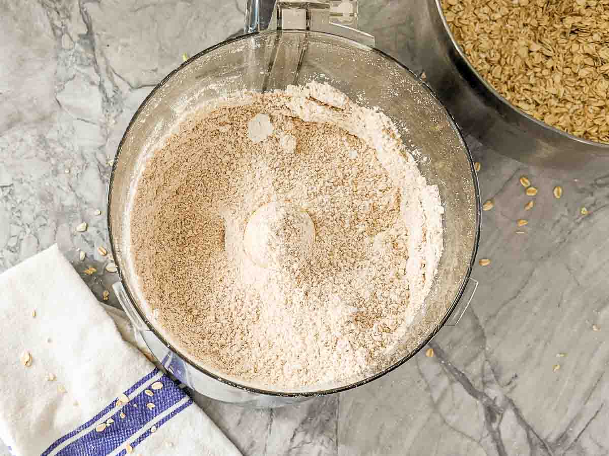 Finished oat flour in a food processor on a marble counter, with a towel and oats in a bowl nearby.
