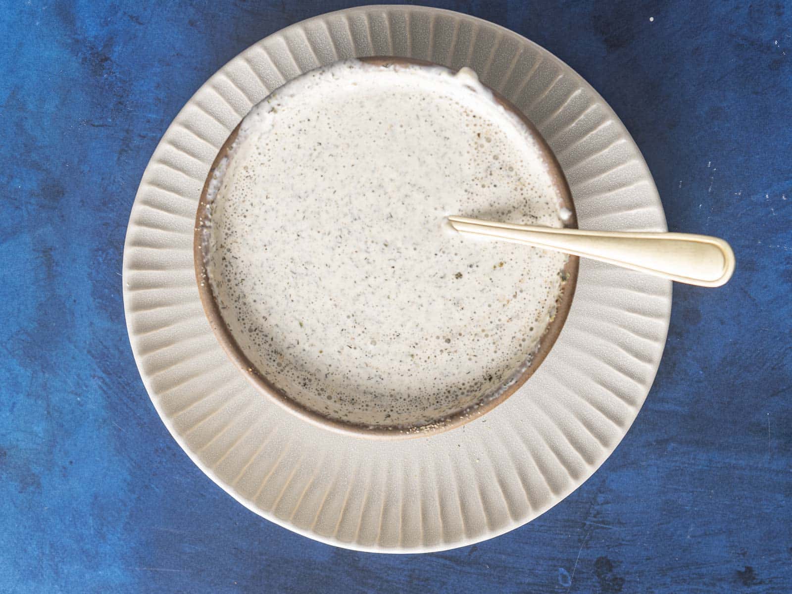 A bowl of creamy garam masala ranch with a spoon rests elegantly on a textured, blue background.