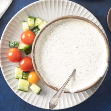 A bowl of creamy Indian salad dressing with a spoon, nestled next to sliced cucumbers and cherry tomatoes on a plate.