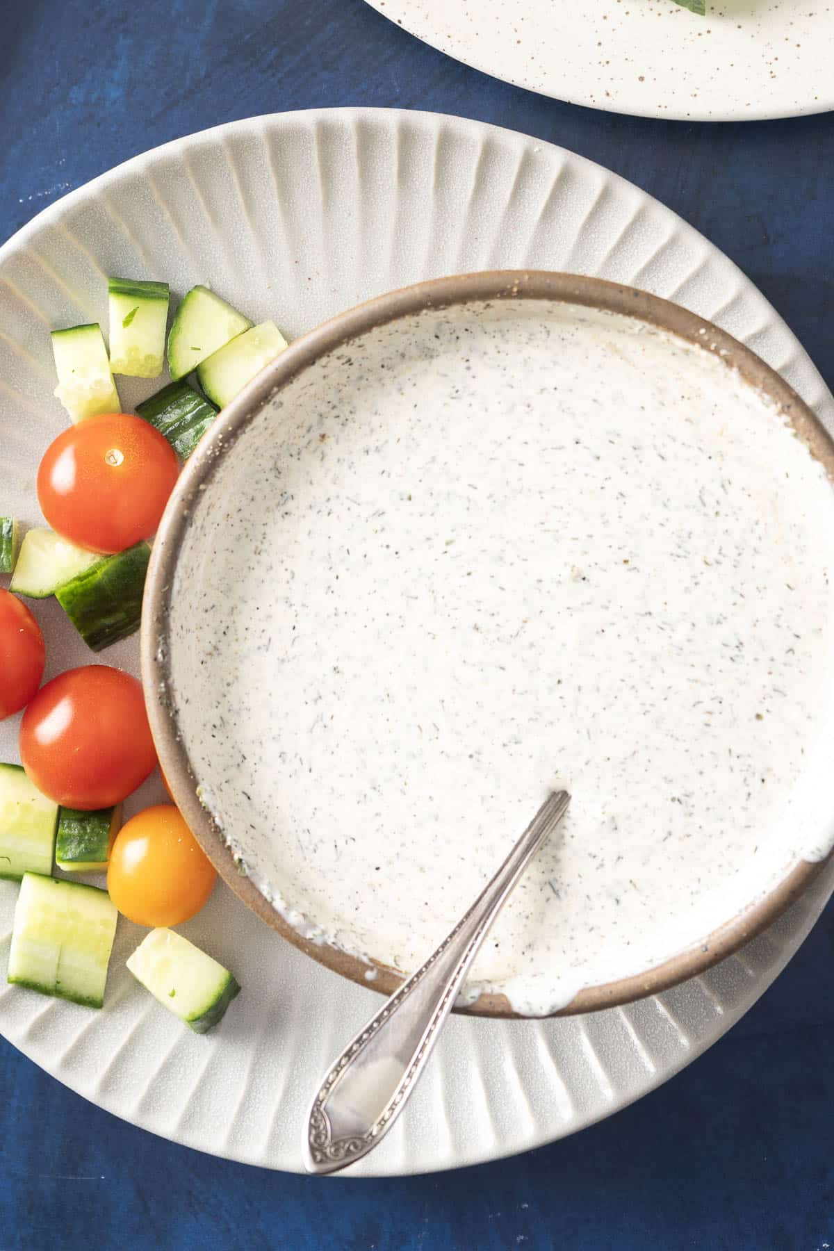 A bowl of creamy Indian salad dressing with a spoon, nestled next to sliced cucumbers and cherry tomatoes on a plate.