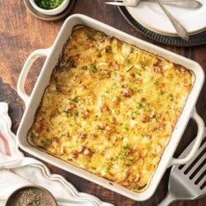 Leek potato gratin, a baked cheesy casserole, sits invitingly in a square dish, garnished with fresh herbs. It's perfectly placed on a wooden table surrounded by plates and utensils, ready to be savored.