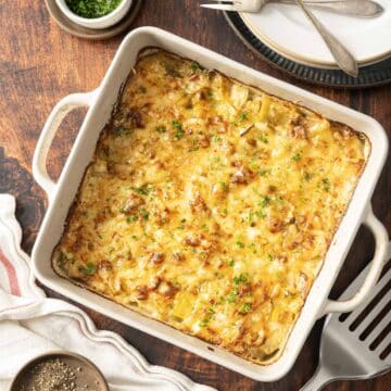 Leek potato gratin, a baked cheesy casserole, sits invitingly in a square dish, garnished with fresh herbs. It's perfectly placed on a wooden table surrounded by plates and utensils, ready to be savored.