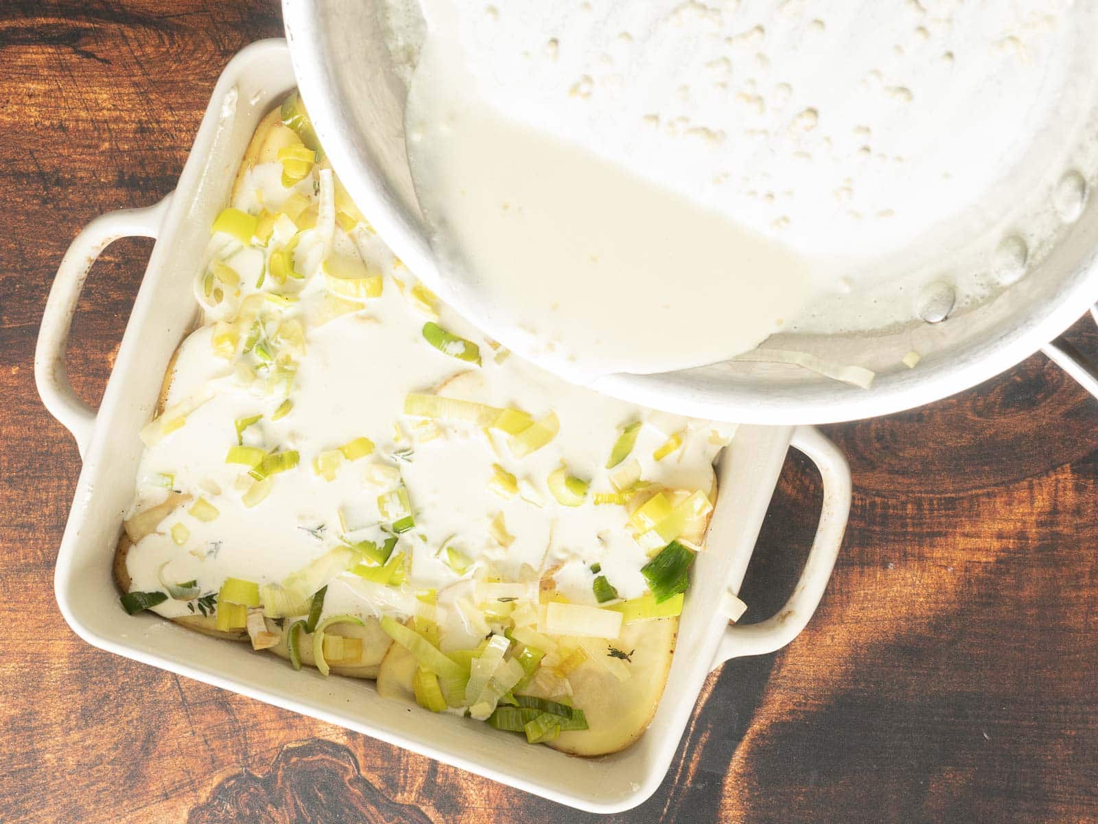 Pouring creamy sauce over sliced potatoes and leeks in a square baking dish, creating a delightful gratin on a wooden surface.