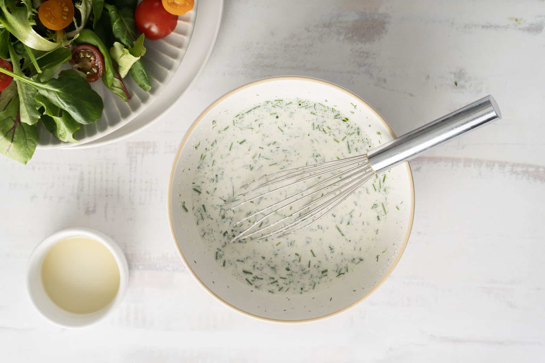 A bowl of creamy buttermilk ranch dressing with a whisk sits beside a vibrant salad and a small cup of dressing on a light surface.