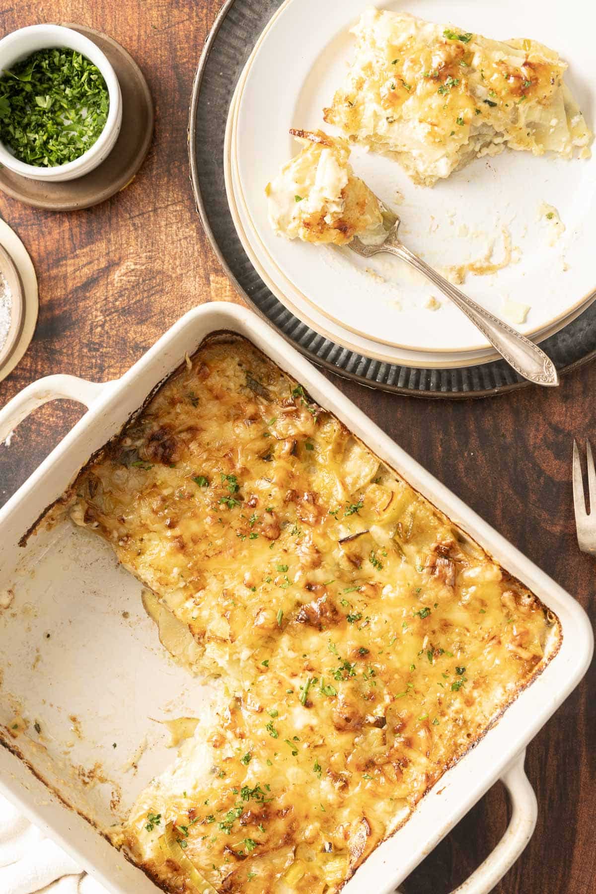 Cheesy lasagna in a baking dish, served on a plate with a fork, garnished with parsley—its rich layers echo the comforting warmth of leek potato gratin.
