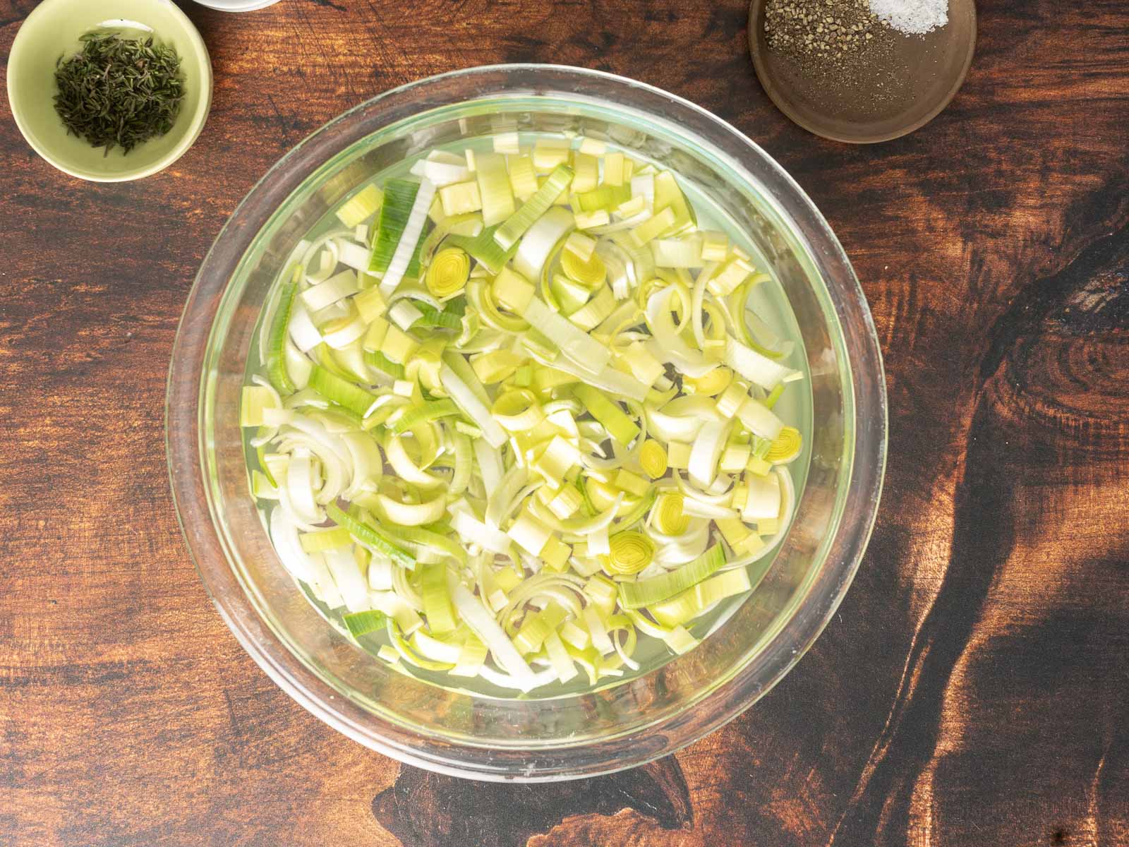 Slices of leeks soaking in a clear glass bowl filled with water on a wooden surface; small bowls of herbs and pepper nearby, perfect for creating a savory leek and potato gratin.