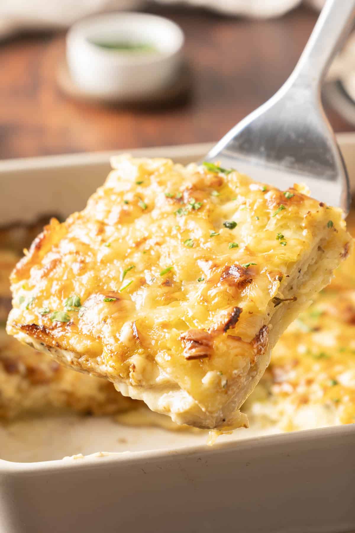 A slice of cheesy baked leek potato gratin with herbs is being served from a dish.