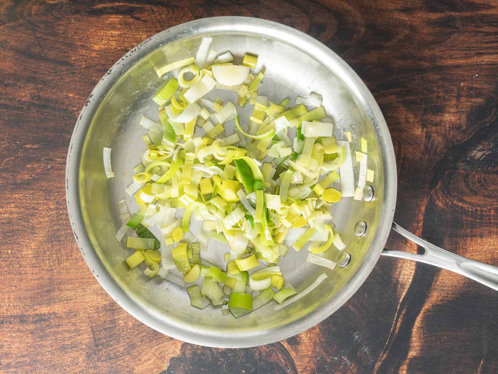 Chopped leeks are being sautéed in a stainless steel pan on a wooden surface, creating the perfect base for a delightful leek and potato gratin.