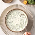 Bowl of creamy herb and buttermilk ranch soup with a spoon, garnished with greens, on a white plate; salad in the background.