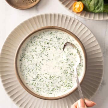 Bowl of creamy herb and buttermilk ranch soup with a spoon, garnished with greens, on a white plate; salad in the background.