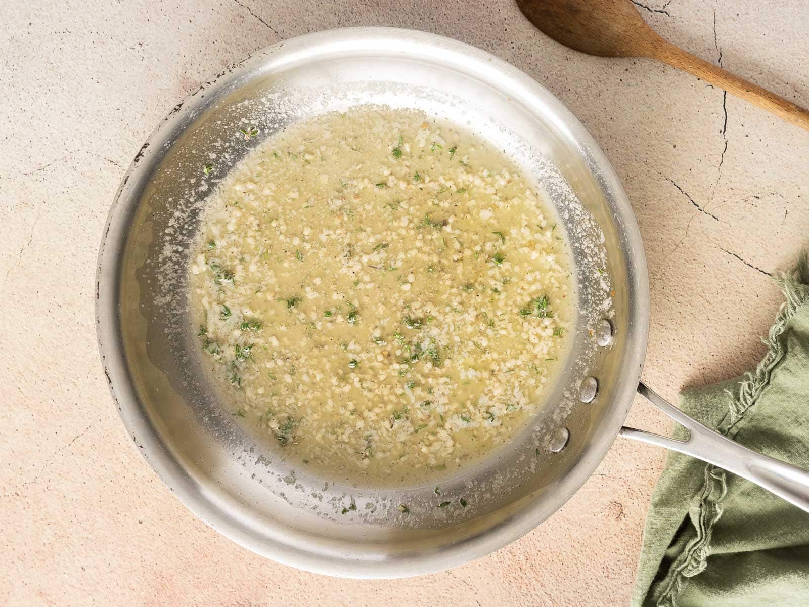 A pan contains a creamy garlic and herb sauce with hints of roasted leeks. A wooden spoon and a green cloth napkin rest nearby on a light-colored surface.
