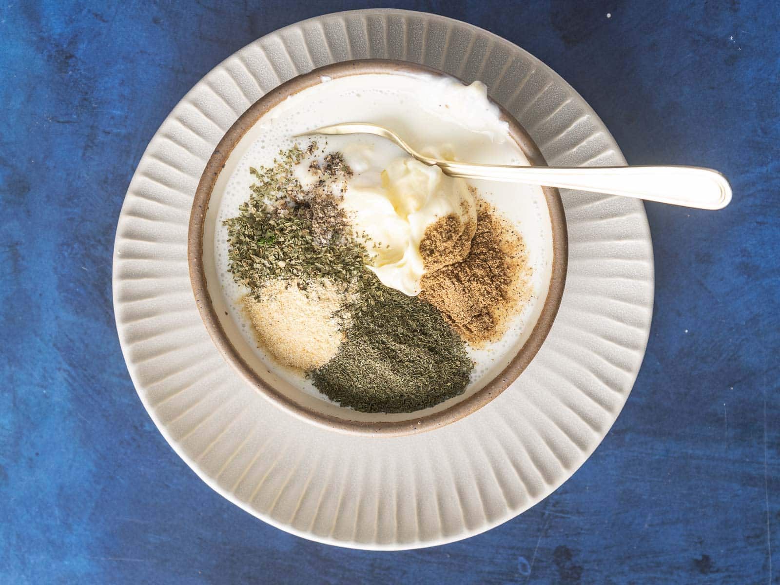 A bowl of buttermilk laced with a medley of herbs and spices, including dill, pepper, garlic, cumin, and a hint of garam masala, on a striking blue background.