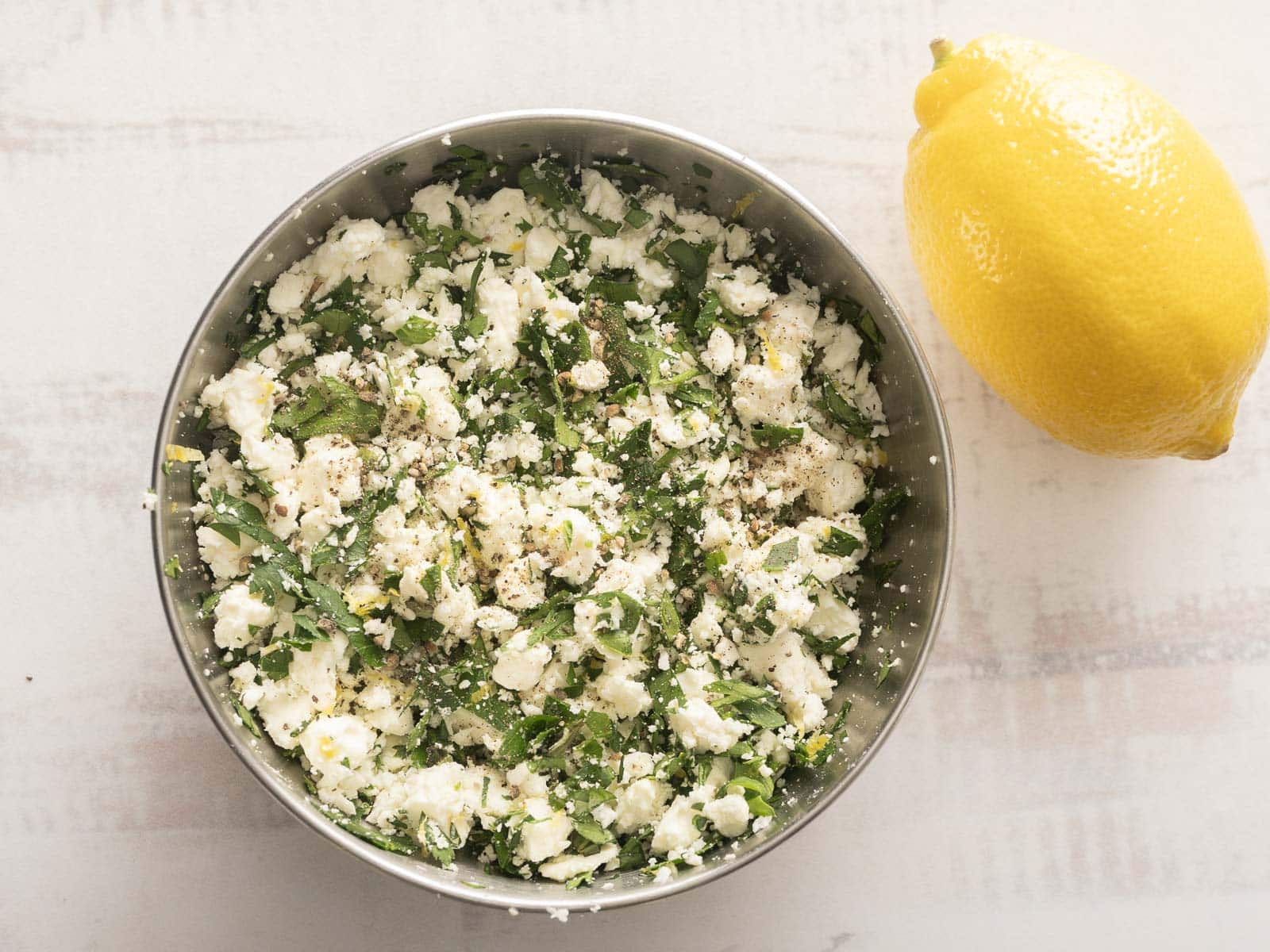A bowl of seasoned feta cheese with herbs stands next to a whole lemon on a light surface.