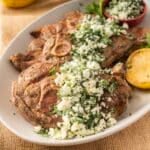 Grilled lamb shoulder chops with crumbled feta gremolata and herbs, artfully garnished with a lemon half and parsley, create a delightful plate.