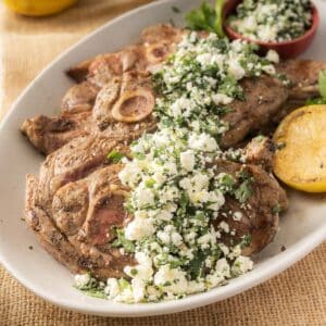 Grilled lamb shoulder chops with crumbled feta gremolata and herbs, artfully garnished with a lemon half and parsley, create a delightful plate.