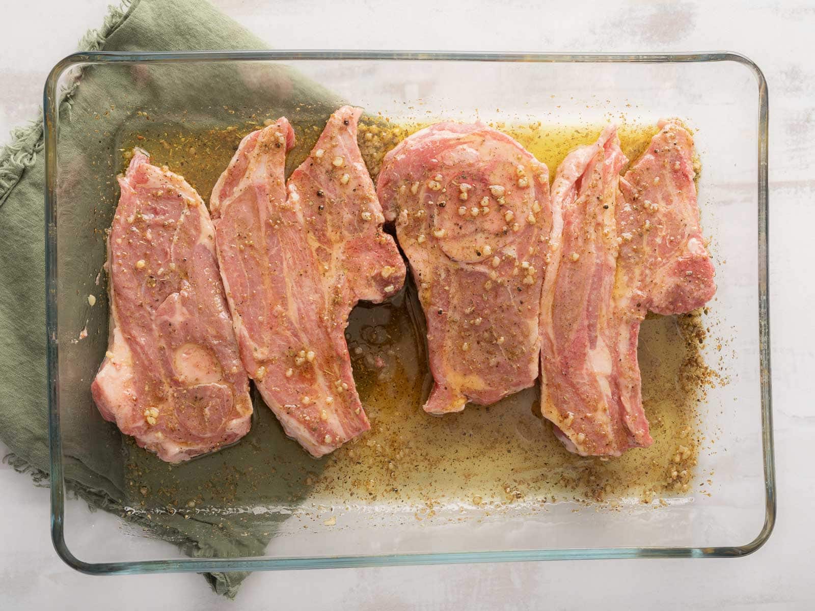 Raw marinated lamb chops in a glass dish rest on a green cloth. 