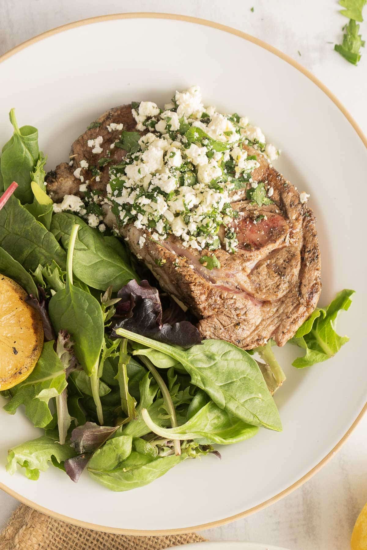 Steak topped with feta gremolata and herbs, served alongside mixed greens and grilled lemon on a white plate.