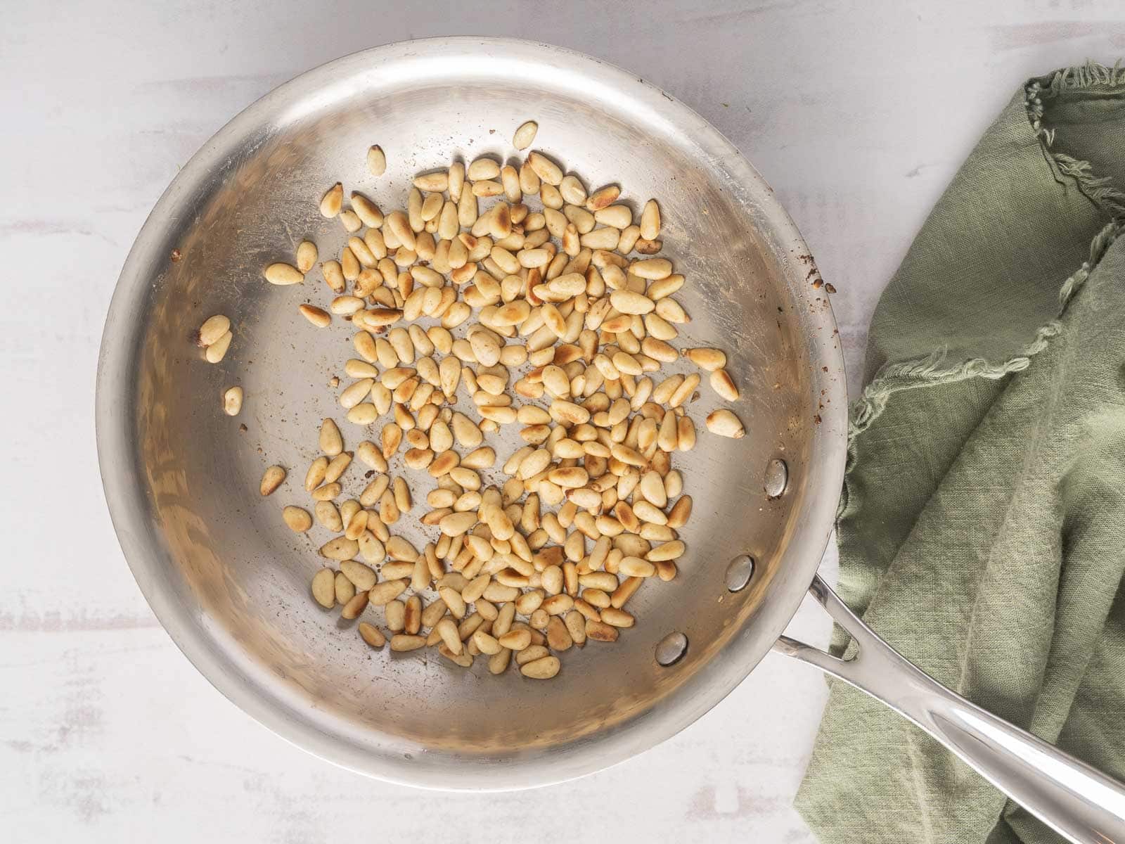Toasted pine nuts in a stainless steel pan next to a green cloth await their turn to add a delightful crunch to your zucchini salad on a light surface.