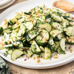 A delightful zucchini salad graces a round white plate, artfully adorned with pine nuts and fresh herbs.