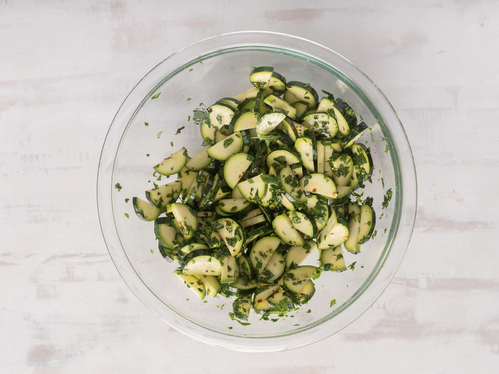 A glass bowl with a refreshing zucchini salad, featuring sliced zucchini tossed in fragrant herbs, rests on a light surface.
