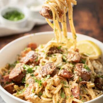 A fork holding creamy spaghetti with sausage and herbs hovers above a bowl, adorned with a lemon and parsley garnish.