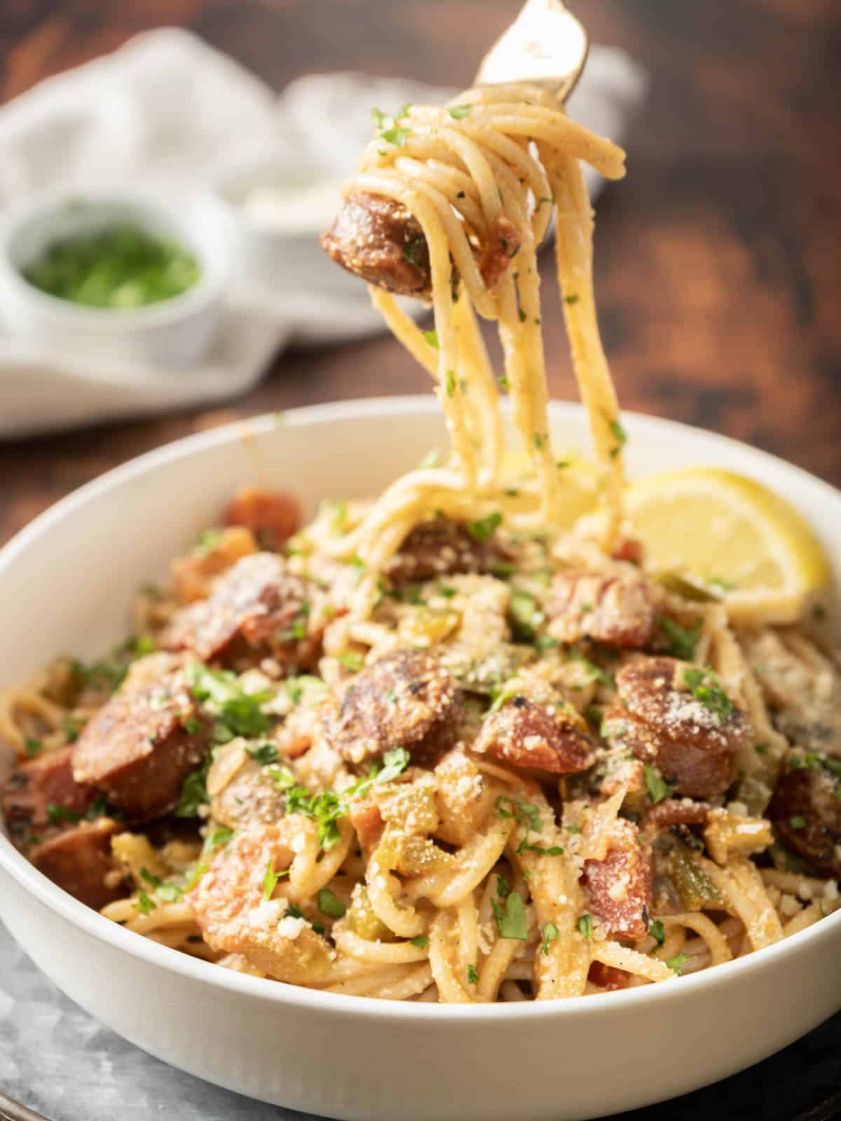 A fork holding creamy spaghetti with sausage and herbs hovers above a bowl, adorned with a lemon and parsley garnish.