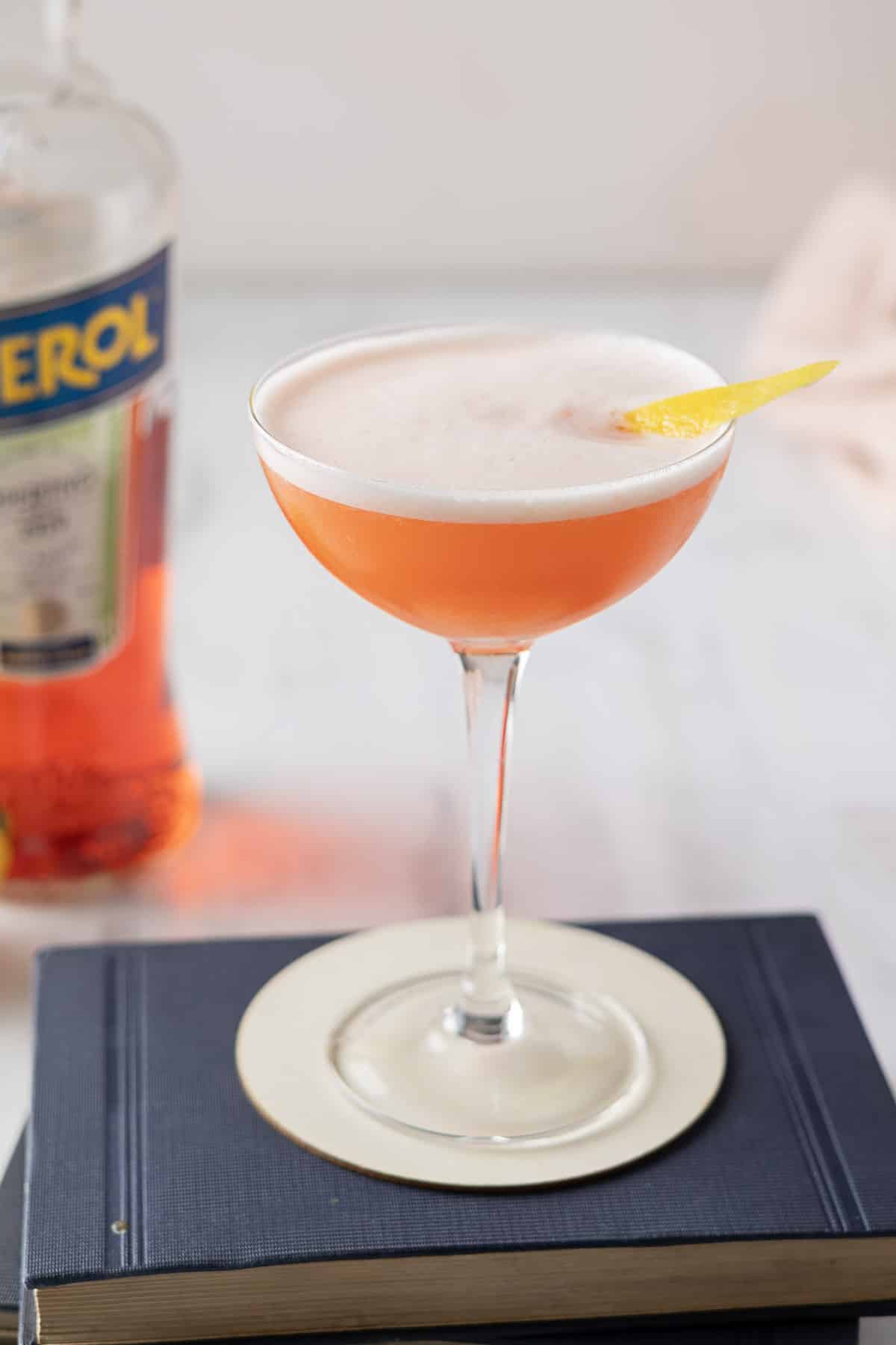 A cocktail in a coupe glass with a lemon twist garnish sits on two stacked books. A bottle of Aperol is in the background.