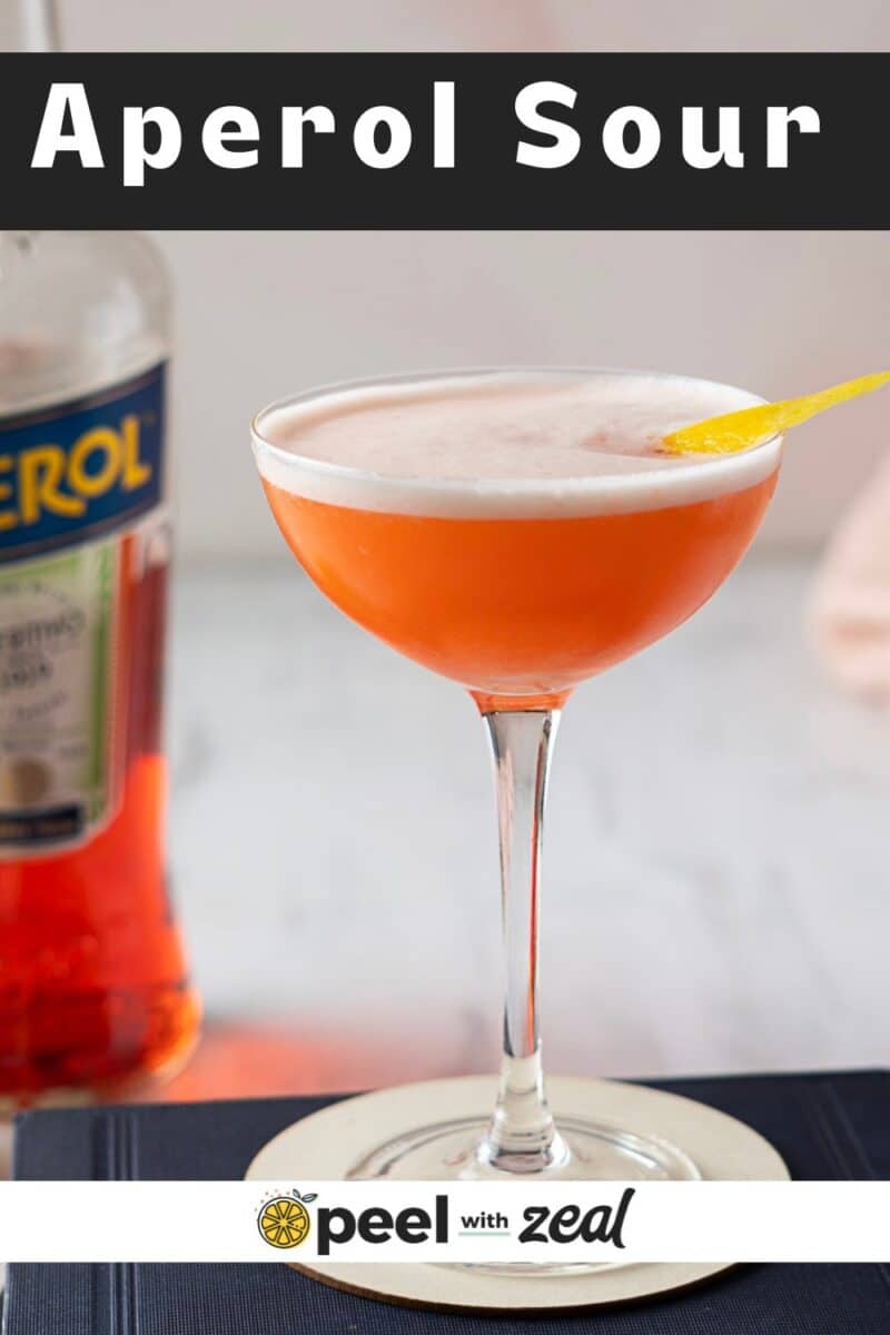 An Aperol Sour cocktail sits elegantly in its glass, garnished with a delicate lemon twist. In the background, a bottle of Aperol watches over this vibrant creation.