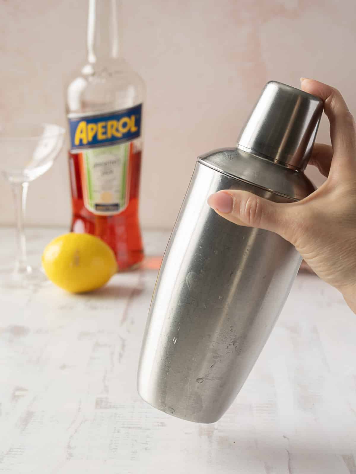 Hand holding a stainless steel cocktail shaker, with a bottle of Aperol, a lemon, and an empty glass on a white surface in the background.