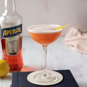 An aperol gin cocktail in a coupe glass with a lemon twist garnish is on a book. A bottle of Aperol and a lemon are in the background.