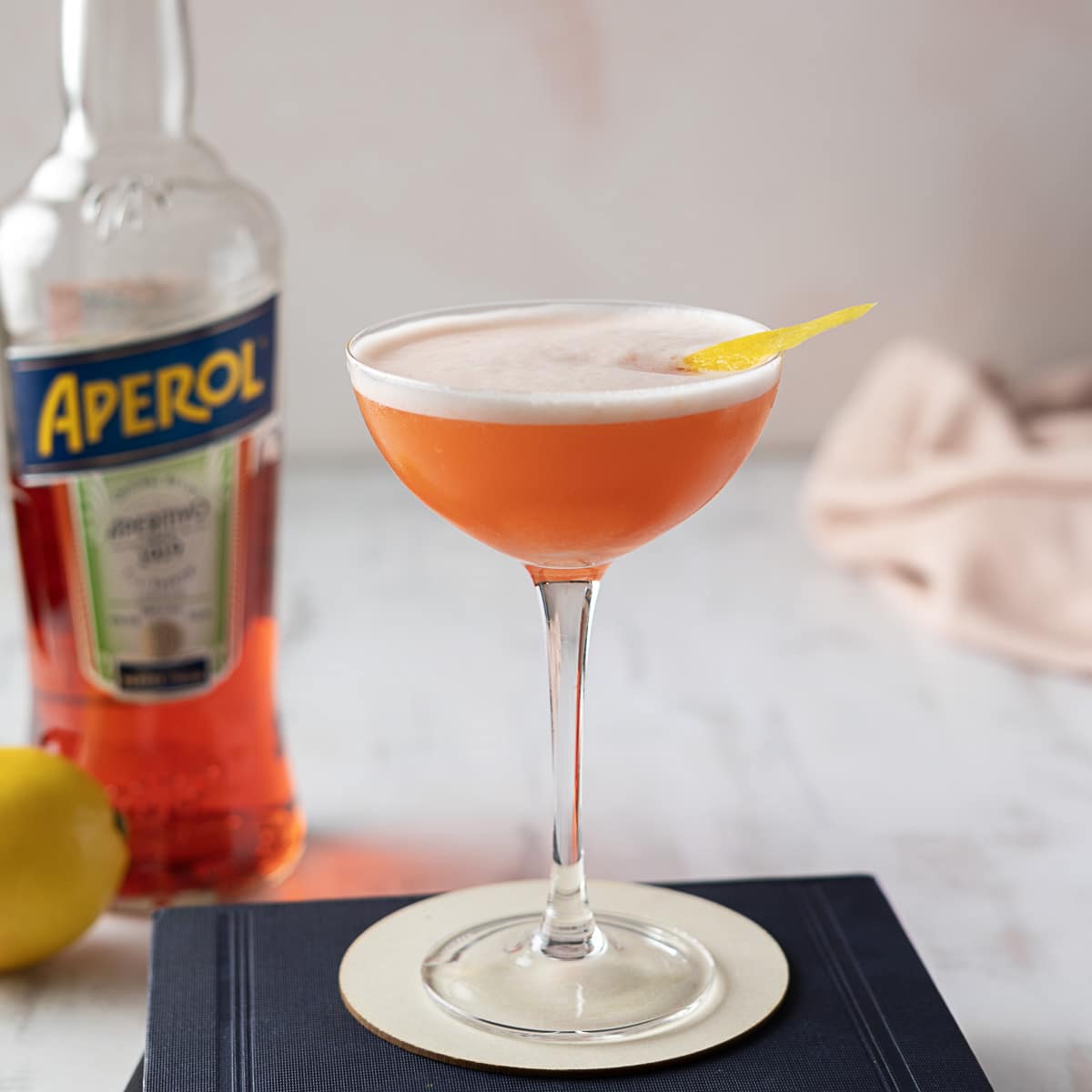aperol sour cocktail in a cocktail glass with a bottle of aperol and lemon on the background
