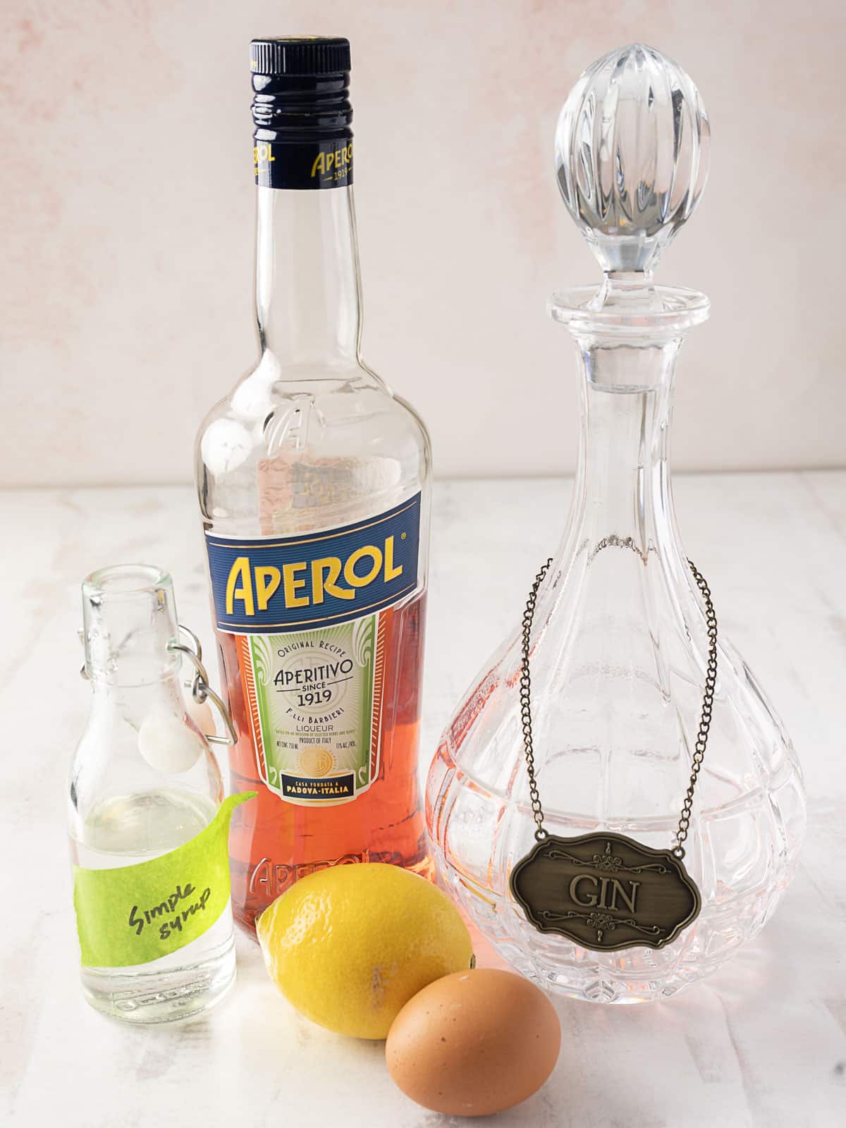 A bottle of Aperol, a gin decanter, a small bottle labeled "simple syrup," a lemon, and an egg are displayed on a table.