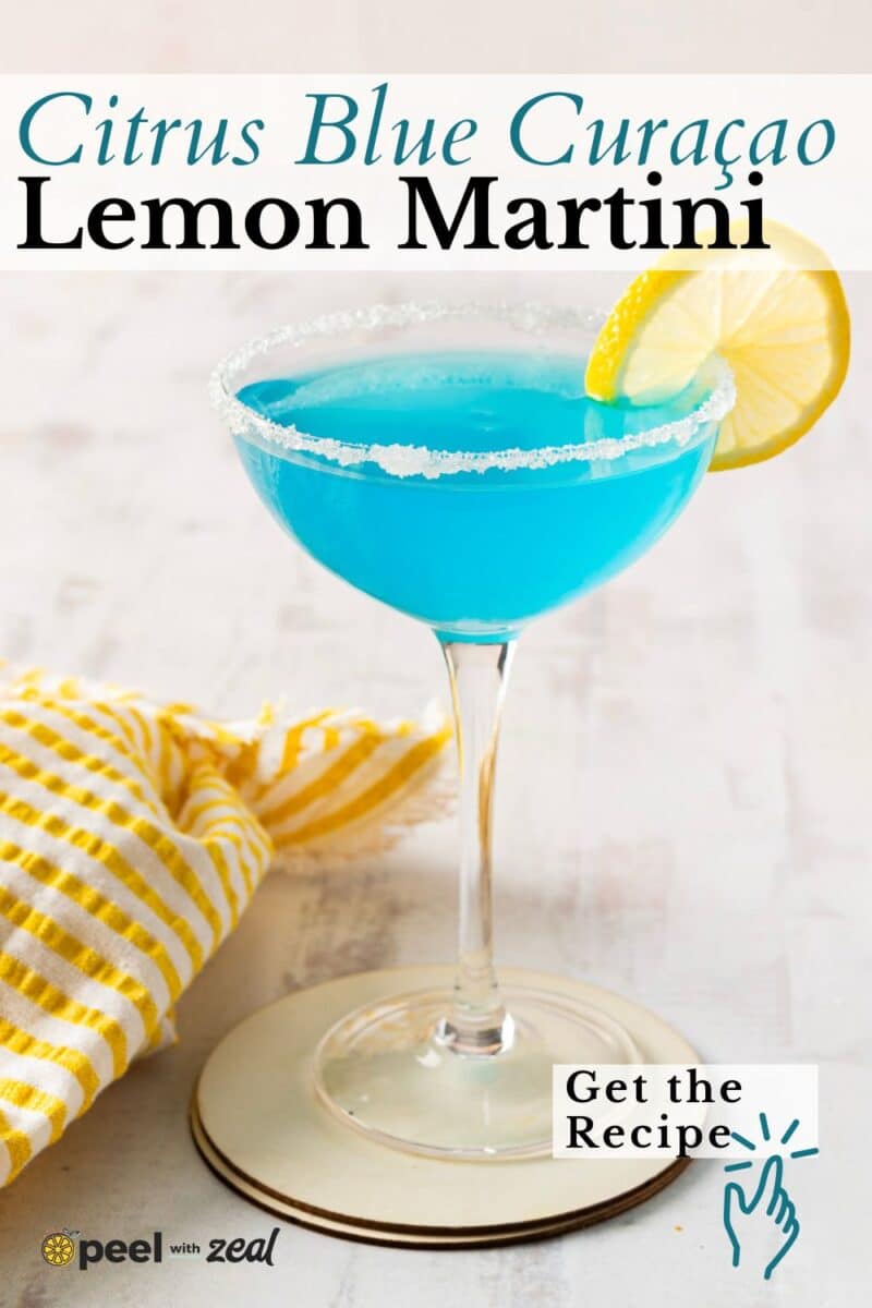 A blue lemon drop martini sits elegantly in a martini glass, its sugar rim sparkling under the light. A lemon slice adorns the glass, perfectly complemented by a yellow-striped cloth beside it.