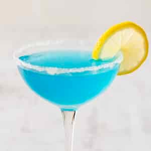 A Blue Lemon Drop Martini dazzles with its vibrant hue, featuring a sugared rim and a lemon slice garnish elegantly perched on the edge of a stemmed glass.