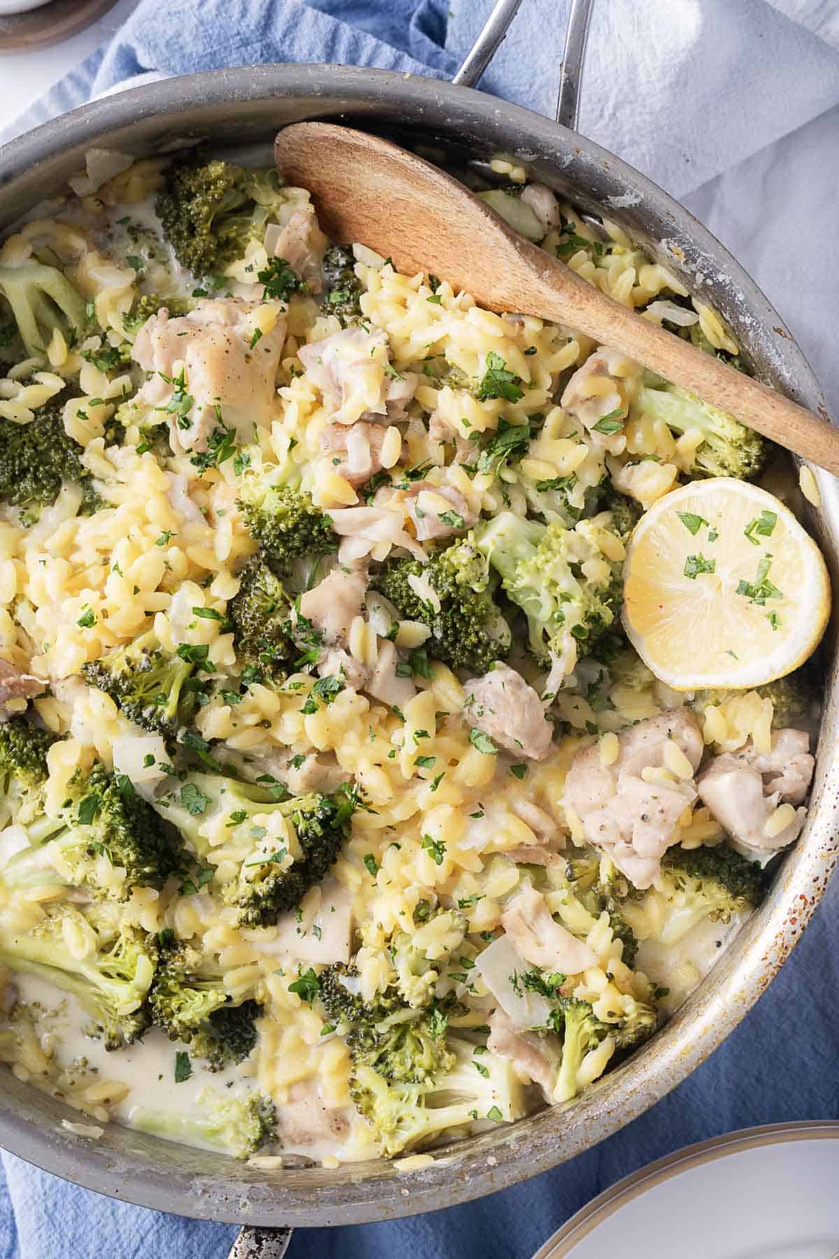 A skillet featuring tender chicken pieces, broccoli, and creamy orzo, all brightened with a lemon slice and garnished with fresh herbs, awaits the delicate touch of a wooden spoon.