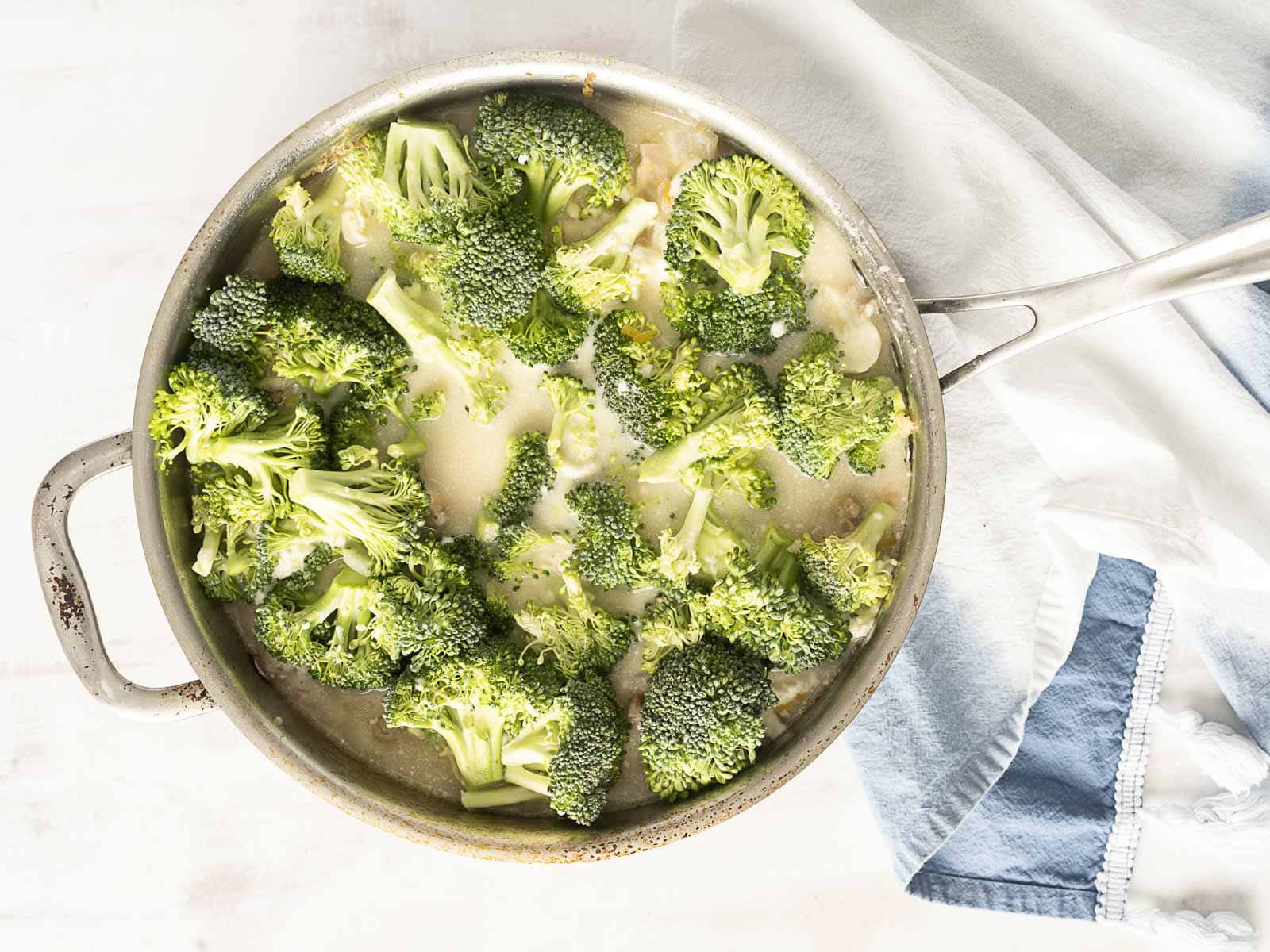 Broccoli cooking in a saucepan on a white surface, with a folded blue and white cloth beside it, creates the perfect setting for whipping up your favorite one-pot meal.
