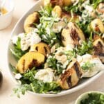A delightful burrata salad featuring grilled peach and arugula, complemented by sliced almonds and a light dressing, elegantly presented on a white oval plate.