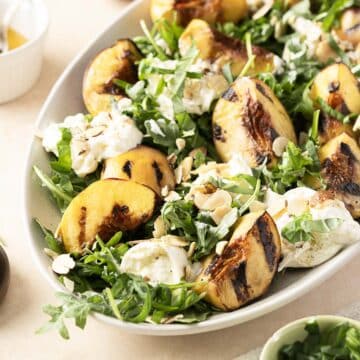 A delightful burrata salad featuring grilled peach and arugula, complemented by sliced almonds and a light dressing, elegantly presented on a white oval plate.