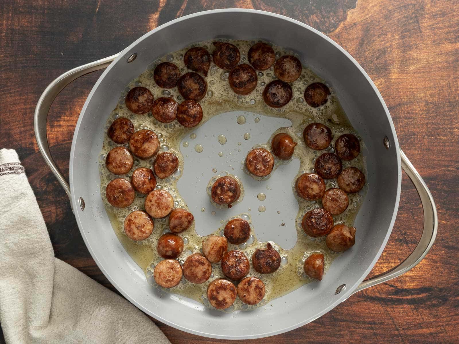 Cajun-spiced sliced sausages sizzle in a pan with oil, set on a wooden surface beside a beige cloth.