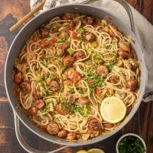 A pan of creamy Cajun-spiced pasta with sausages, mushrooms, tomatoes, and herbs, garnished with lemon slices and fresh parsley.
