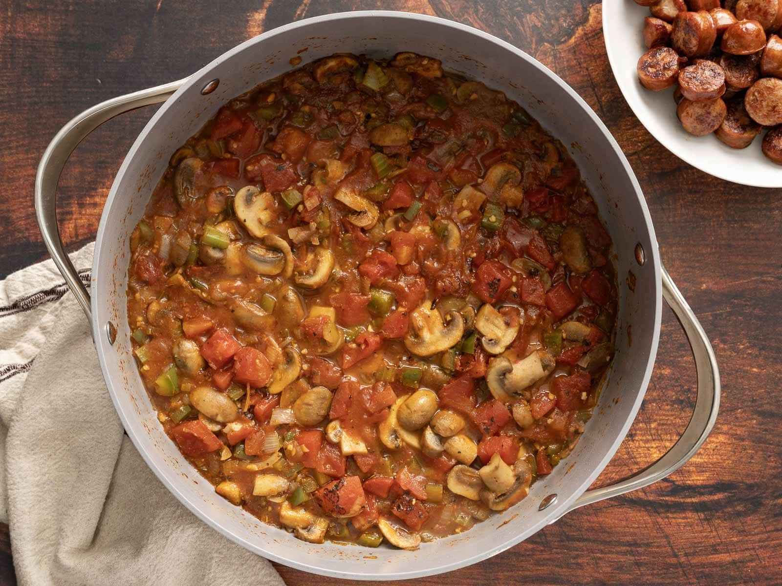 A pot of tomato, mushroom, and vegetable stew sits on a wooden surface alongside a plate of Cajun-spiced sausages.