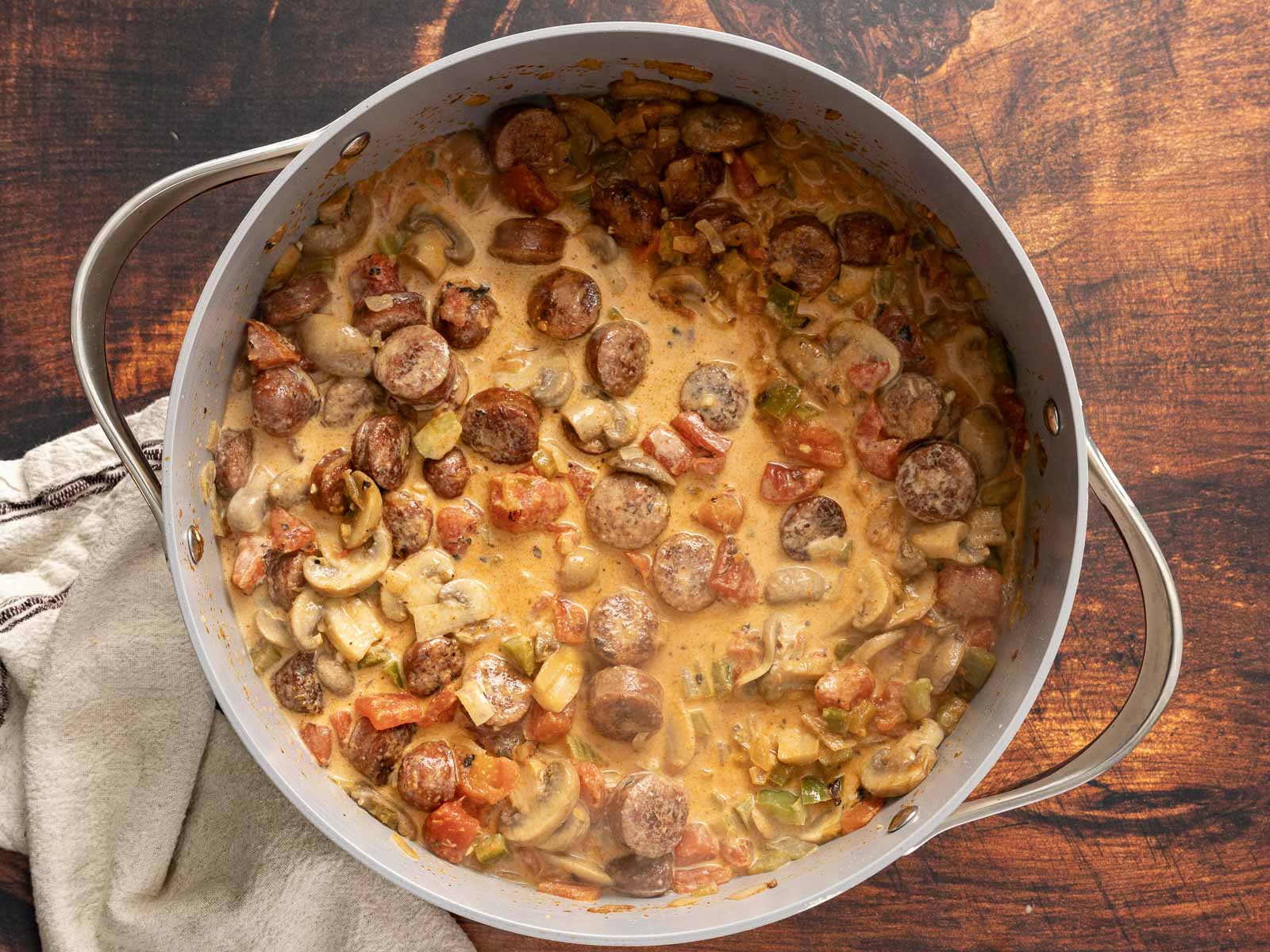 A large pot of creamy Cajun sausage, mushroom, and vegetable stew sits on a wooden table, with a cloth beside it.