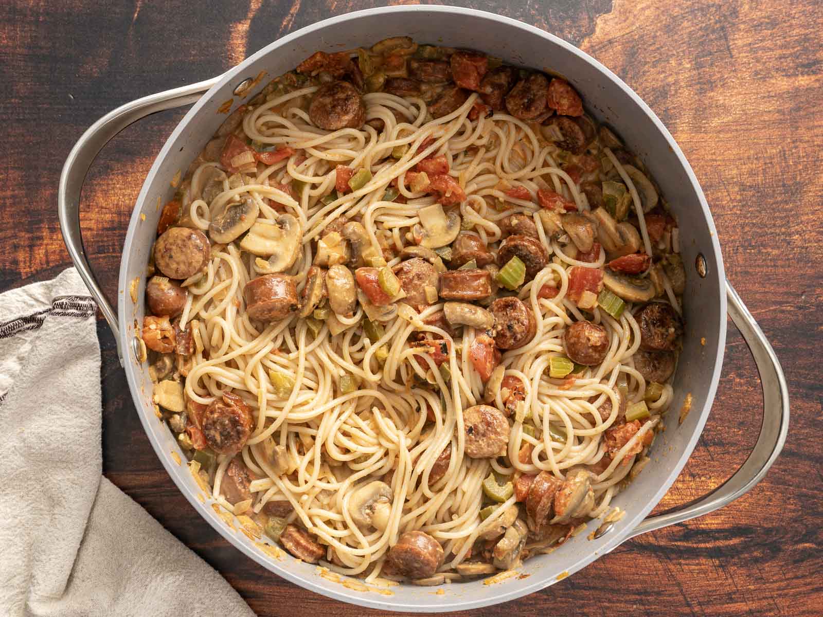 Cajun pasta with sausage, mushrooms, and tomatoes sits simmering in a large pot on a wooden surface, next to a cloth.