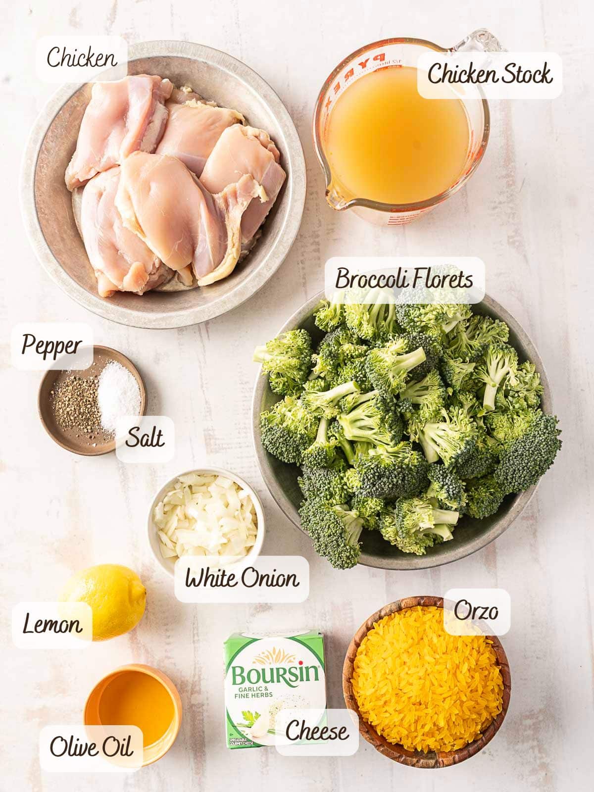 Ingredients for a Chicken Orzo Bake are arranged on a surface: chicken, chicken stock, broccoli florets, orzo, cheese, olive oil, lemon, chopped onion, salt, and pepper.