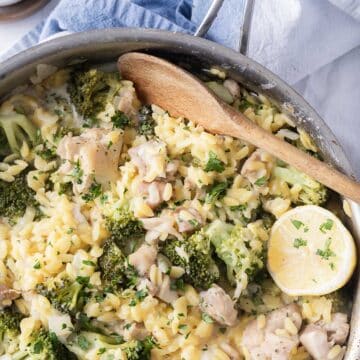A one-pot chicken orzo bake with creamy Boursin, featuring vibrant broccoli and fresh lemon slices, garnished with chopped herbs and served with a wooden spoon.